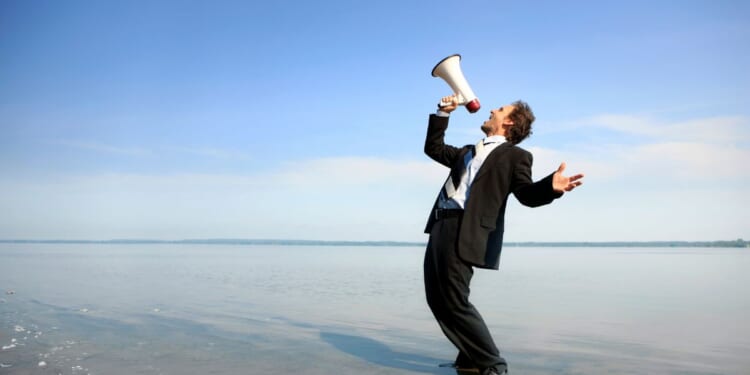 This stock image shows a businessman yelling at the sky with a megaphone.