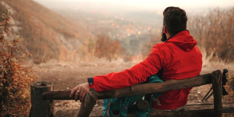 This photo shows a hiker sitting alone on an outdoor bench with his backpack and hiking poles next to him. The spot where he is sitting overlooks city lights in the distance.