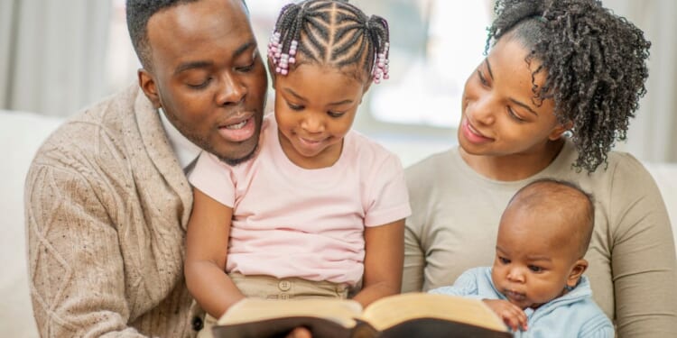 A family of four are sitting on the couch together, reading a Bible story.