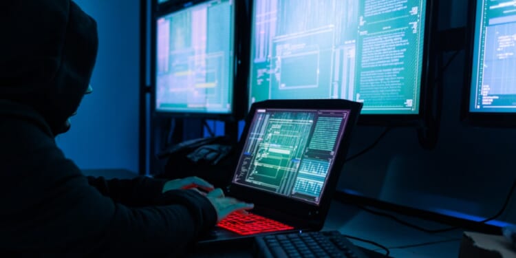 A hooded figure with a laptop sits before a bank of computer screens.