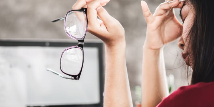 In this image a woman appears to be struggling with eye strain as she removes her glasses due to tiredness from computer screen work.