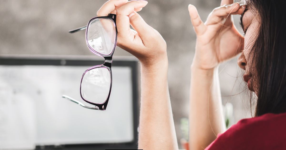 In this image a woman appears to be struggling with eye strain as she removes her glasses due to tiredness from computer screen work.