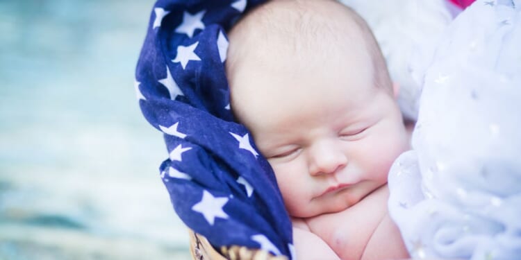 This image shows a newborn baby sleeping in a basket filled with patriotic scarves.