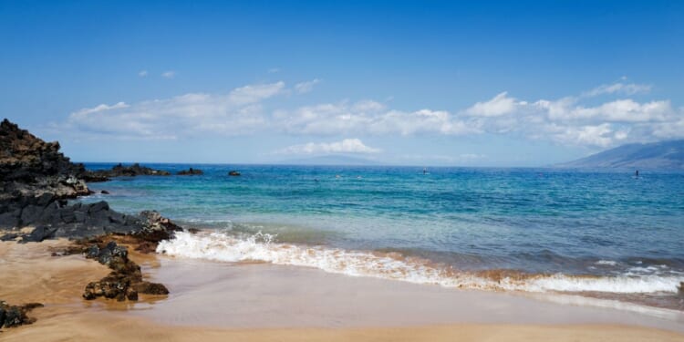 On a sunny day at Wailea beach, small waves break on the sand near volcanic rocks as tourists snorkel and use stand up paddleboards, Wailea, Maui, Hawaii, 2016.