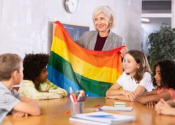 In a classroom, a woman holds up a rainbow colored flag.