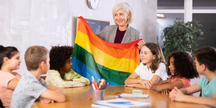 In a classroom, a woman holds up a rainbow colored flag.
