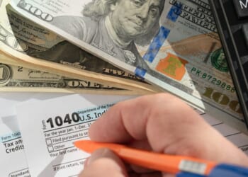 A close-up of the hand of a person filling out a form 1040 on a desk with some dollar bills and a calculator.