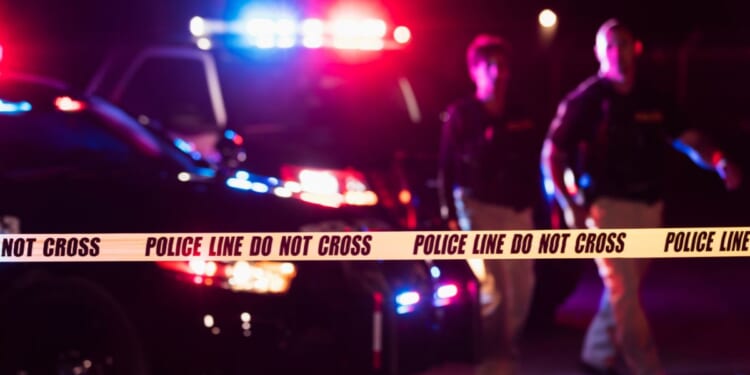 Two police officers walk in front of flashing patrol cars at a crime scene.
