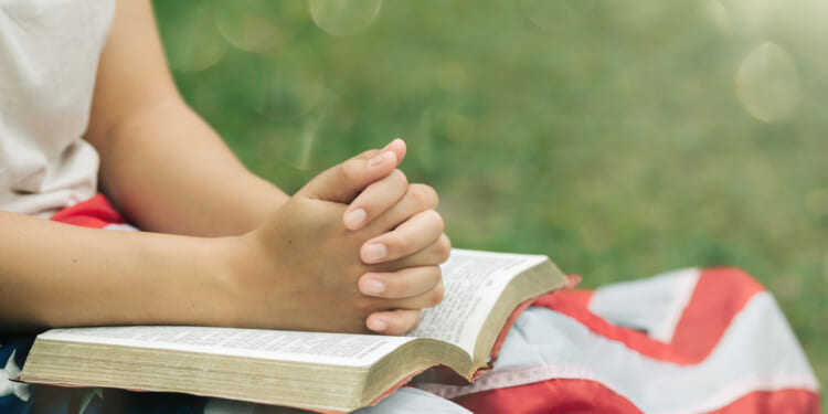 Close-up of young person with flag and Bible on their lap in outdoor setting.