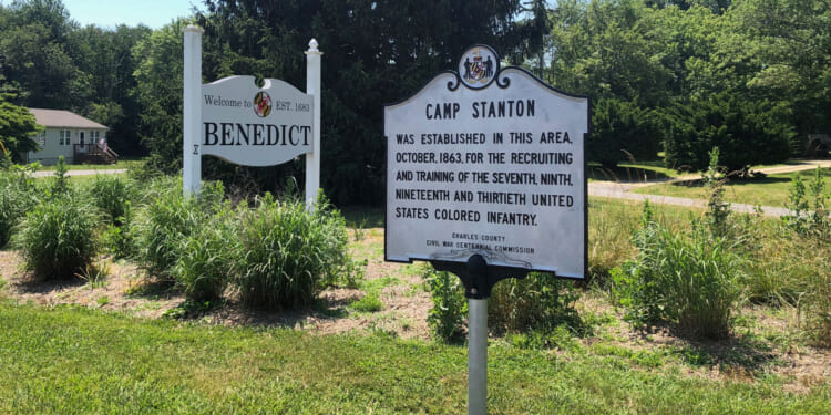 A grey historic marker for "Camp Stanton" alongside a welcome sign for Benedict.