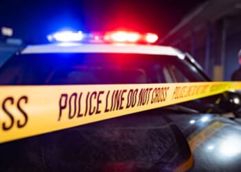 A police car flashes its lights as an office unfurls a line of exclusionary tape to cover the area.