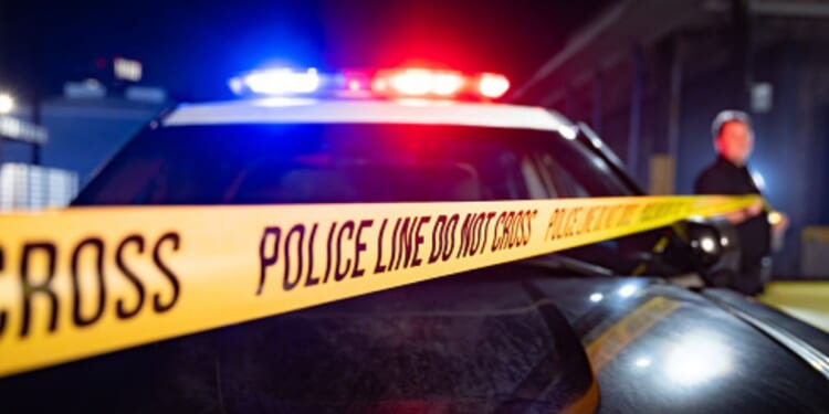A police car flashes its lights as an office unfurls a line of exclusionary tape to cover the area.