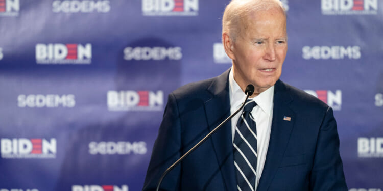 Former President Joe Biden stands at a microphone, with a banner featuring his name and that of "SCDEMS."