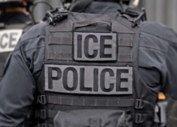 An ICE officer in his uniform stands among his colleagues.