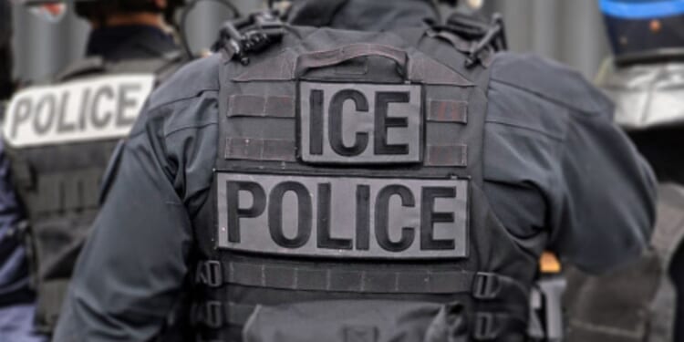 An ICE officer in his uniform stands among his colleagues.
