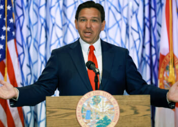 Ron DeSantis behind a podium with his mouth open and arms stretched wide.