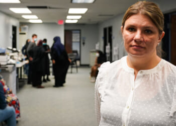 Aimee Bock, in a white blouse, stands in a room with others in the background.
