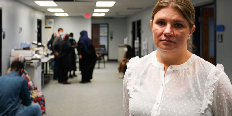 Aimee Bock, in a white blouse, stands in a room with others in the background.