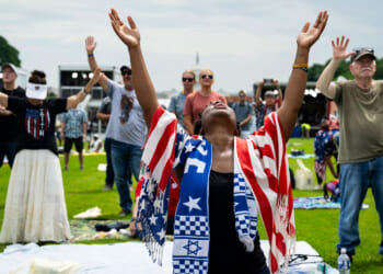 A Black woman draped in American flag and Israeli scarf lifts hands to heaven, with US Capitol in distant background.
