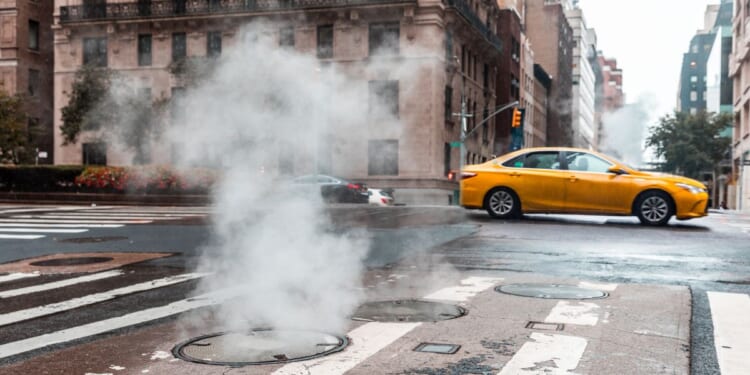 An urban street with steam rising from a manhole and a taxi in motion in New York.