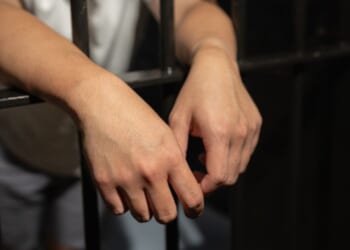 A man stands in a cell and leans over the bars.