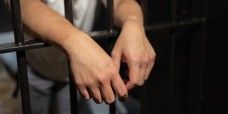 A man stands in a cell and leans over the bars.