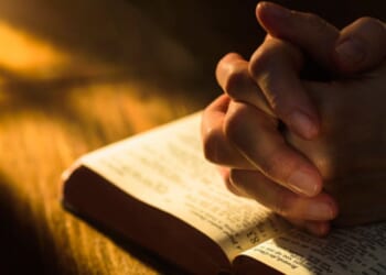Hands clasped in a prayerful position over an open Bible.