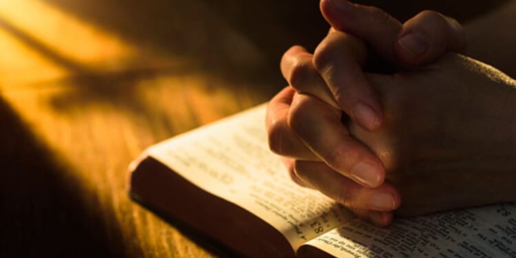 Hands clasped in a prayerful position over an open Bible.