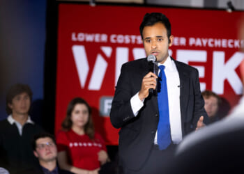 Republican Vivek Ramaswamy speaks in front of a crowd with a microphone in his hand and his red campaign backdrop behind him.