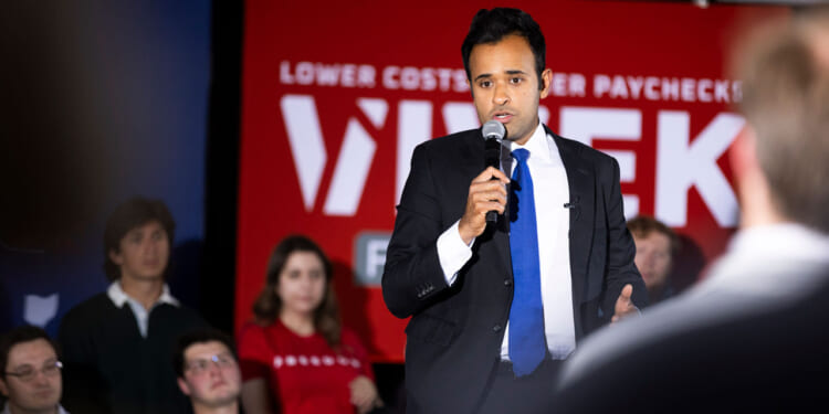 Republican Vivek Ramaswamy speaks in front of a crowd with a microphone in his hand and his red campaign backdrop behind him.