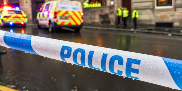 Blue-on-white police tape across a street with ambulance and officers in background.
