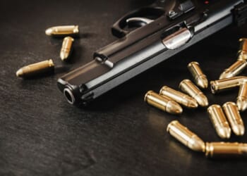 A gun lays on a table near dozens of bullets.