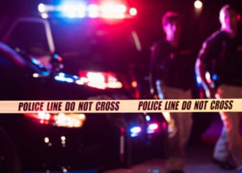 Police lights flash at a crime scene as two officers walk by.