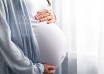 A pregnant woman holds her stomach as she looks out a mirror.