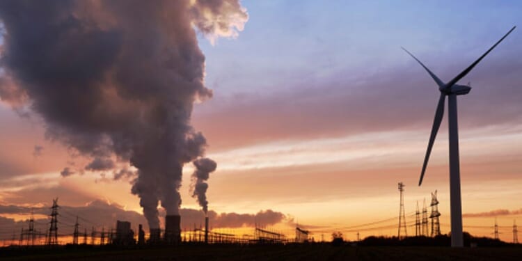 A wind turbine turns as factories pump out smoke in the evening.