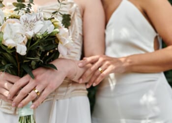 Two women dressed as brides stand close to each other as one embraces the other.