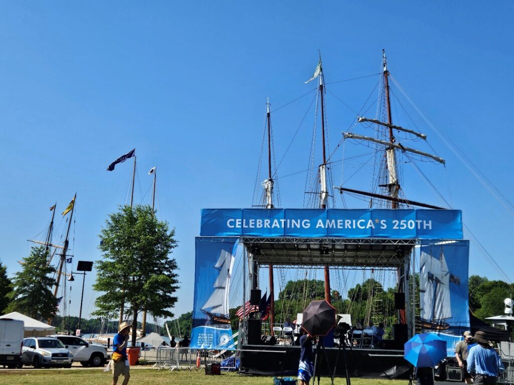 An outdoor stage with banner "Celebrating America's 250" with the masts of three tall ships rising in the background