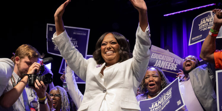 Janeese Lewis George in a white suit waves with both hands in the air as supporters celebrate behind her.