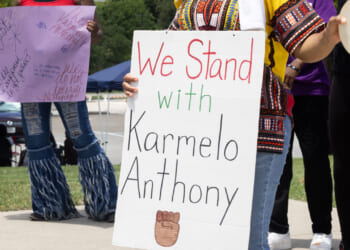 Protester in African garb holds sign reading "We Stand With Karmelo Anthony."