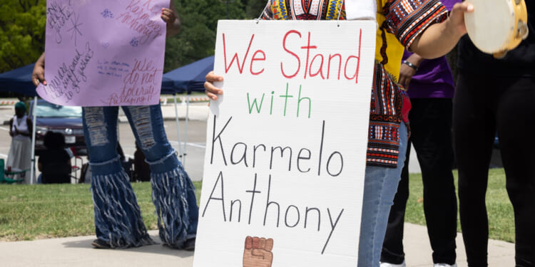 Protester in African garb holds sign reading "We Stand With Karmelo Anthony."