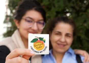 Two women are pictured, while one of them holds a Georgia voter sticker in front of a tree.