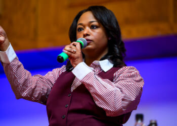 Janeese Lewis George in a maroon vest and maroon and white striped shirt holds a microphone to her mouth and has her right hand stretched out and clenched.