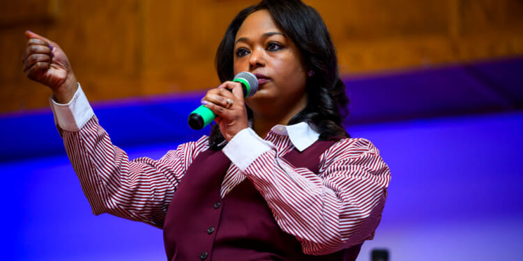 Janeese Lewis George in a maroon vest and maroon and white striped shirt holds a microphone to her mouth and has her right hand stretched out and clenched.