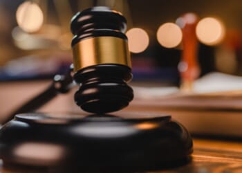 A gavel sits on a wooden block as a balancing scale and book are pictured behind.