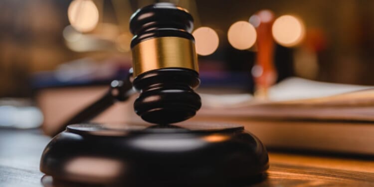 A gavel sits on a wooden block as a balancing scale and book are pictured behind.