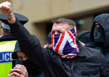 A protester, his face covered in a UK flag, raises his fist as other protesters and a police officer stand behind him.