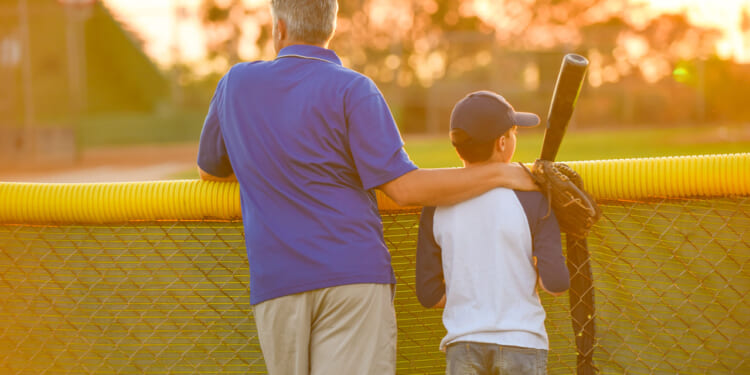 A father in a blue shirt has his arm around his son, who's holding a bat and wearing a baseball jersey and cap. The pair is looking over a ball field in late afternoon.