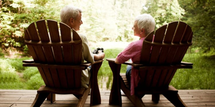 The rear view of a senior couple relaxing in Adirondack chairs on a wooden deck.