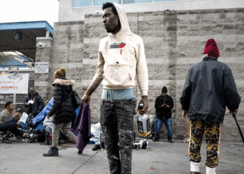 A young man in a hoodie and ripped jeans stands on a city street, looking to the side.