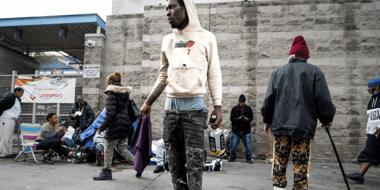 A young man in a hoodie and ripped jeans stands on a city street, looking to the side.